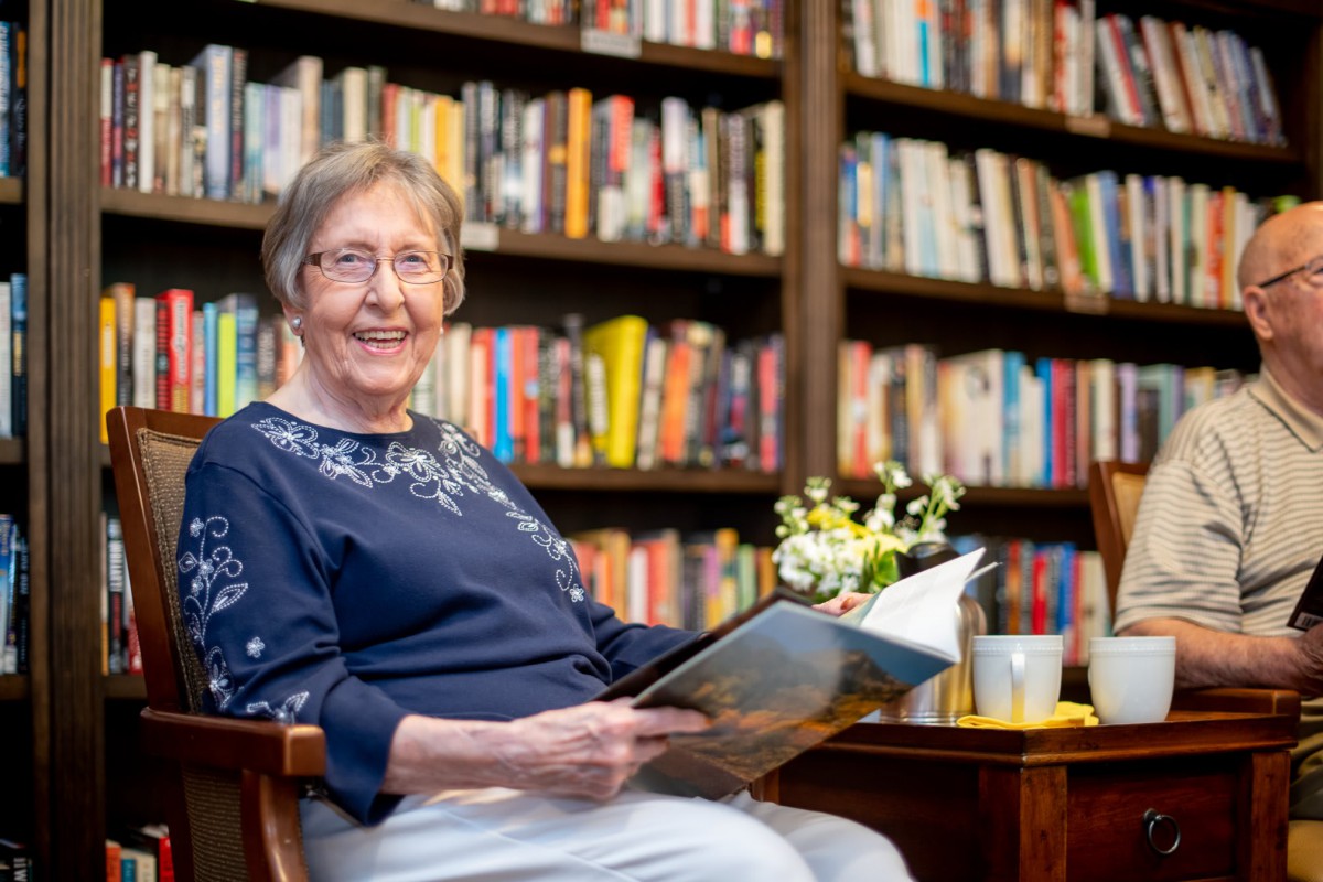 An elderly woman sits comfortably in a library, enjoying a book with a cup of tea and flowers nearby.