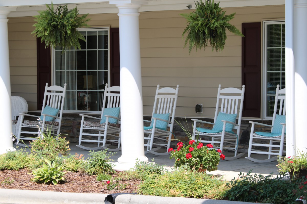 A row of white rocking chairs on a porch adorned with hanging plants and flowers.