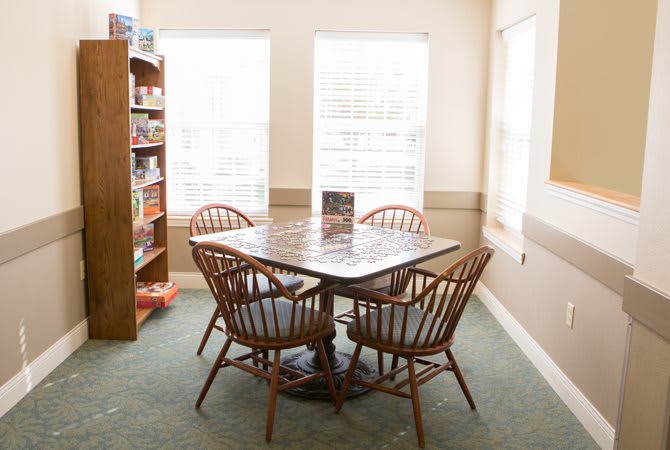 A cozy dining area with a wooden table and chairs, surrounded by natural light from the windows and a nearby bookshelf stocked with board games.