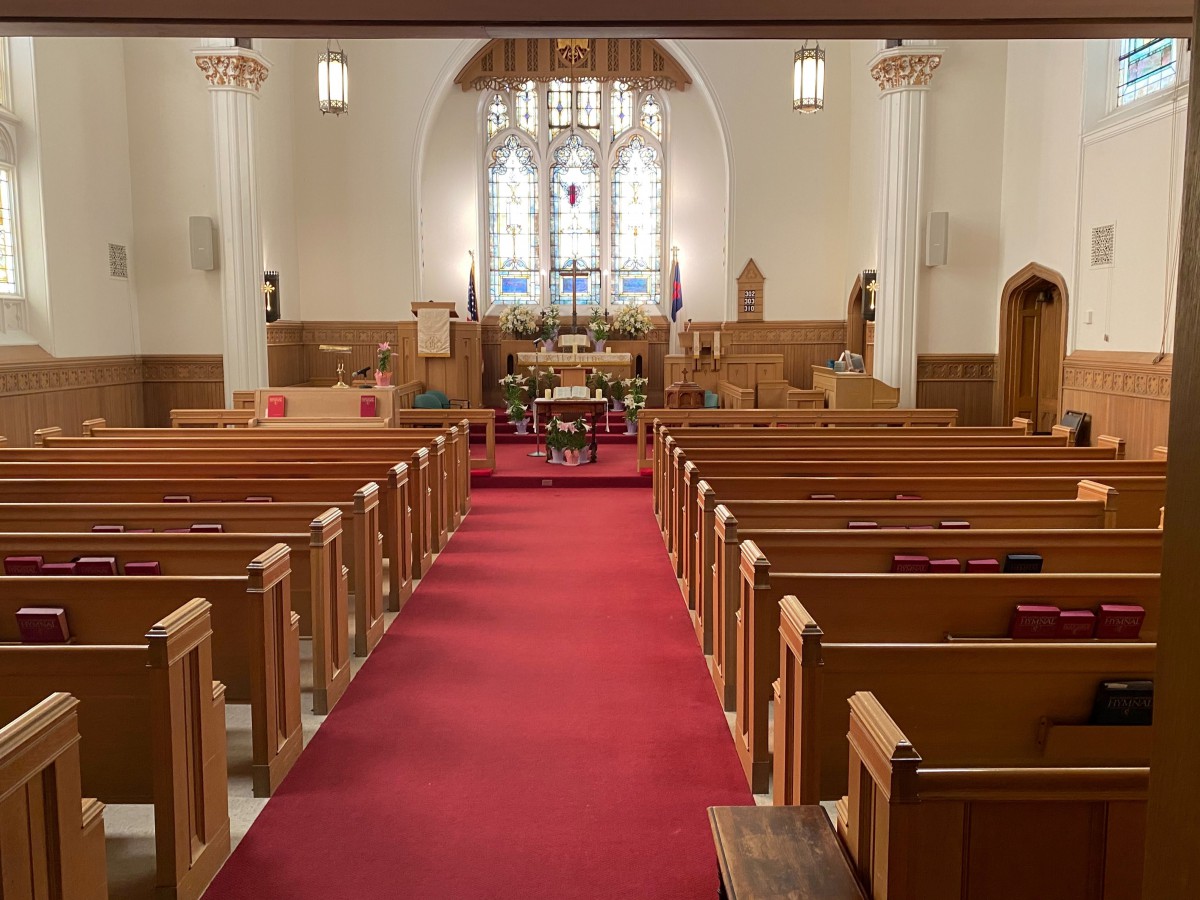 Interior view of a church with wooden pews, red carpet, and a stained glass window behind the altar.