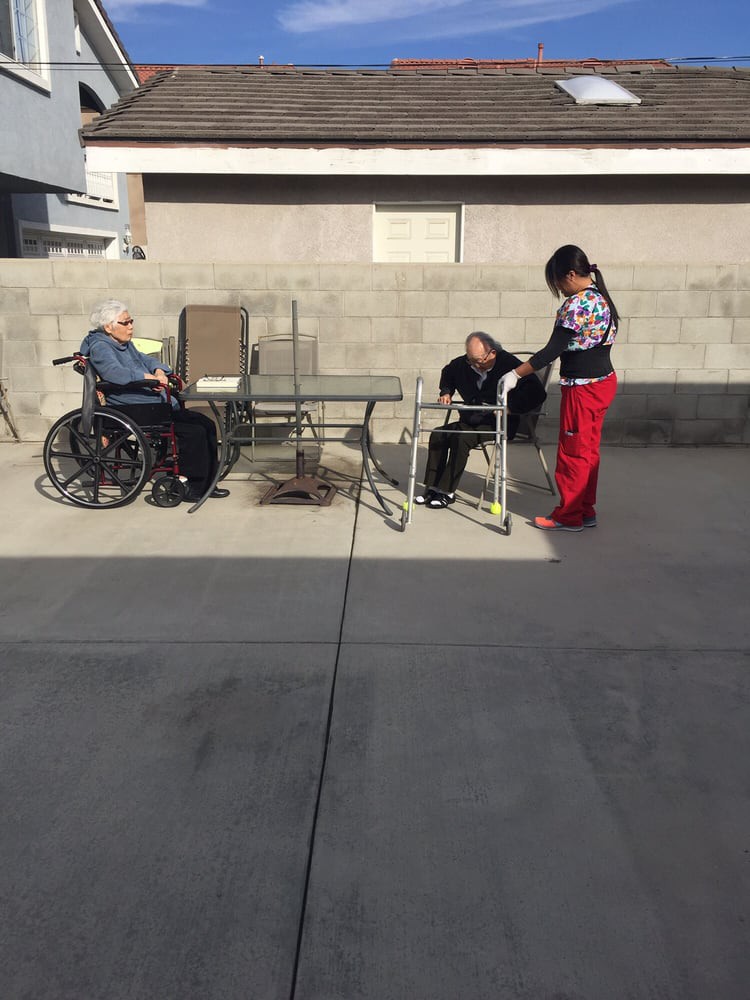 Two elderly individuals are sitting outdoors, one in a wheelchair and the other with a walker, accompanied by a caregiver on a sunny concrete patio.