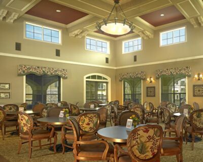 A spacious and elegantly decorated dining area with multiple tables and chairs, featuring large windows and a central chandelier.