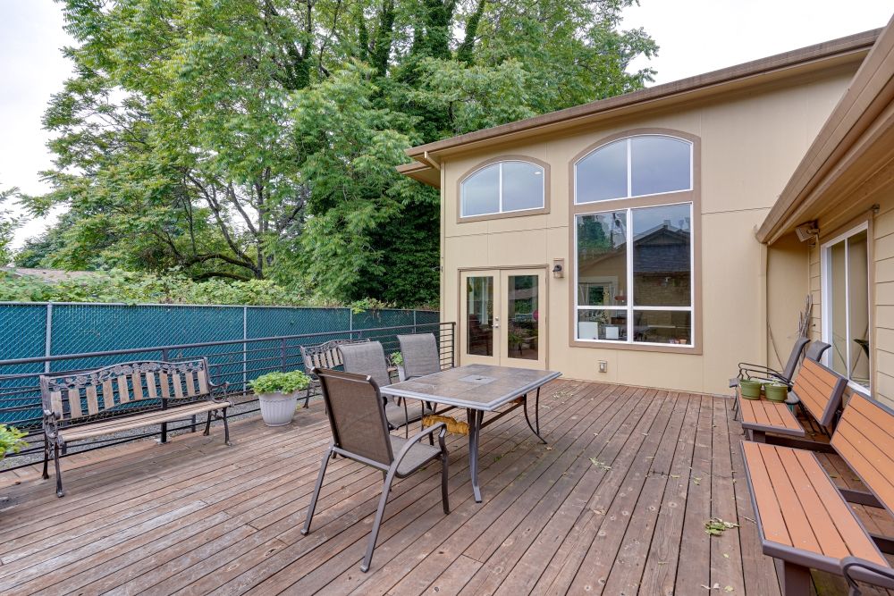 A spacious outdoor patio with a wooden deck, featuring a table and chairs, adjacent to a modern house surrounded by greenery.