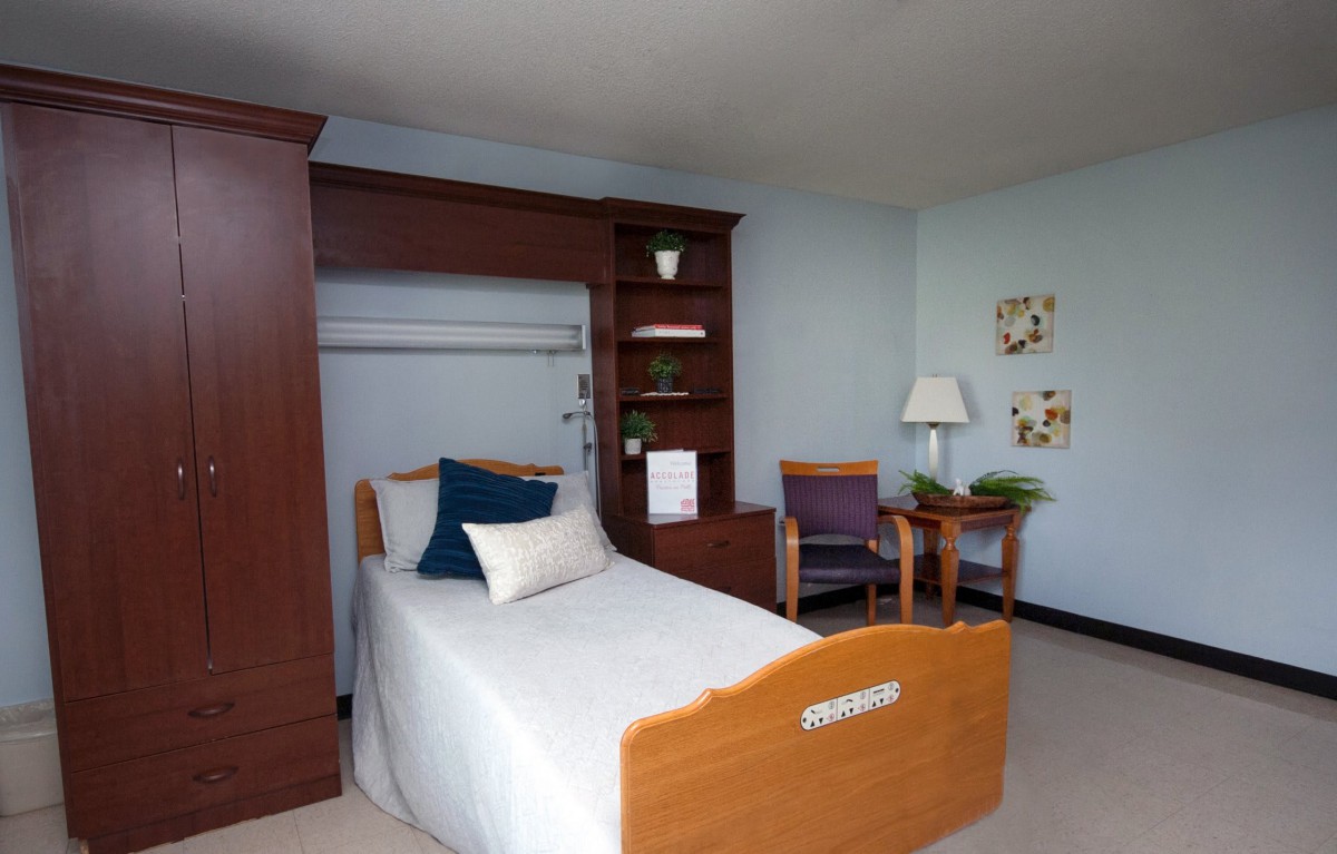 A simple hospital room with a single bed, wooden furniture, and minimal decor.