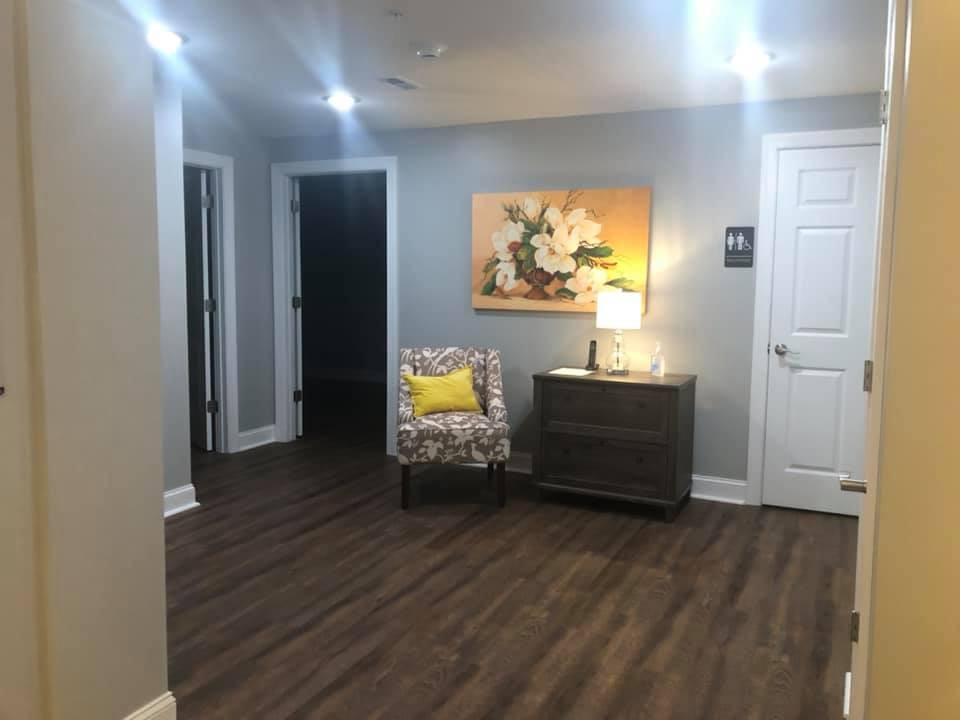 A well-lit hallway with wooden flooring, featuring a floral-patterned chair, a small table with a lamp, and a decorative painting on the wall.