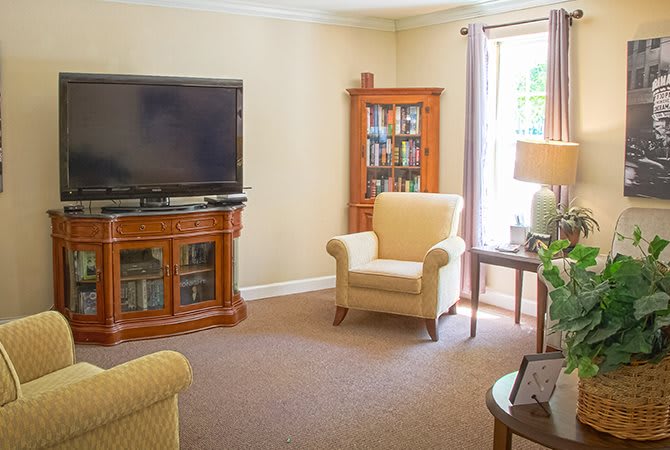 A cozy living room with a television, an armchair, and a bookshelf near a window.