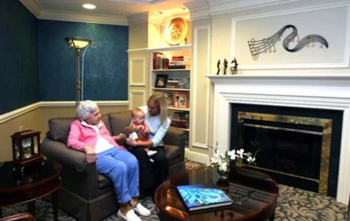A cozy living room scene with a family gathering near the fireplace, featuring an elderly person and a baby.