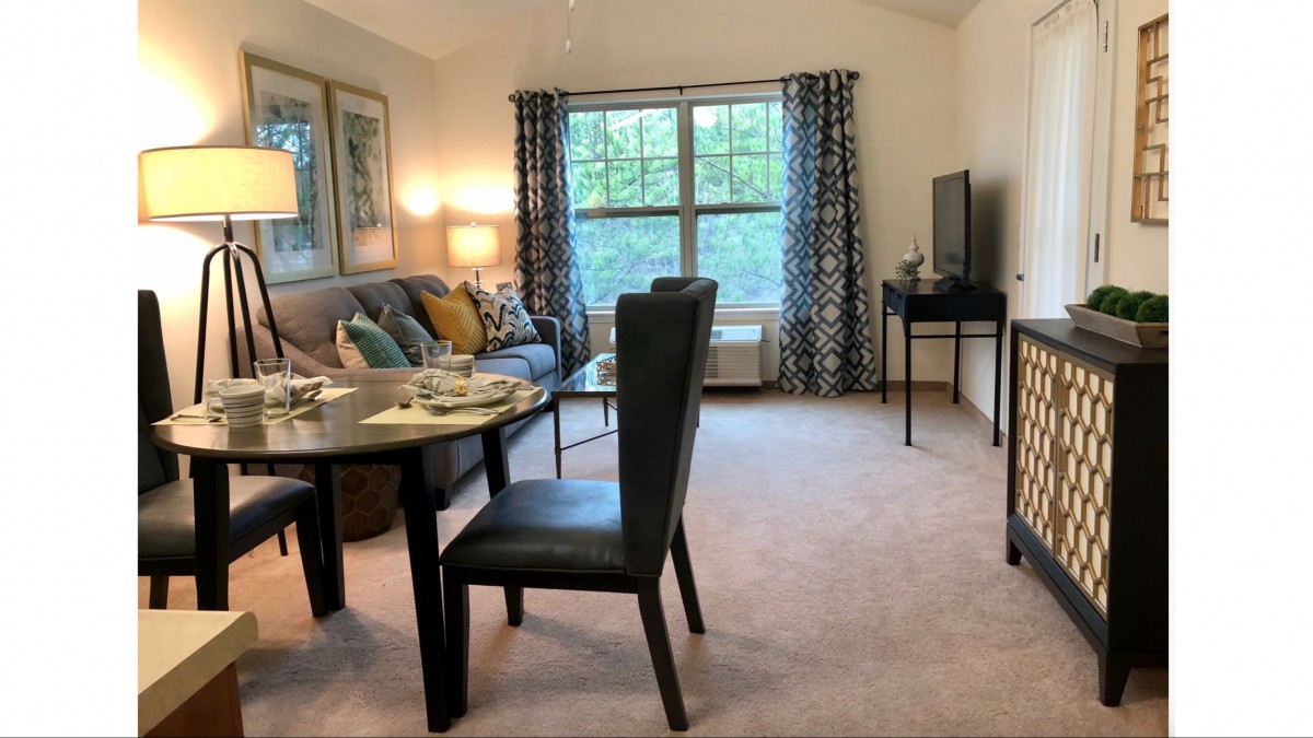 A cozy living room with a dining area, featuring a sofa, dining table set, and a television beside a large window.