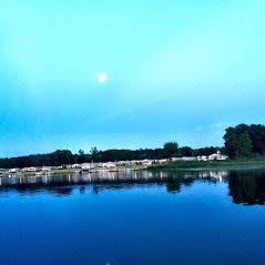 A serene view of a lake at dusk with the moon rising and its reflection on the water.
