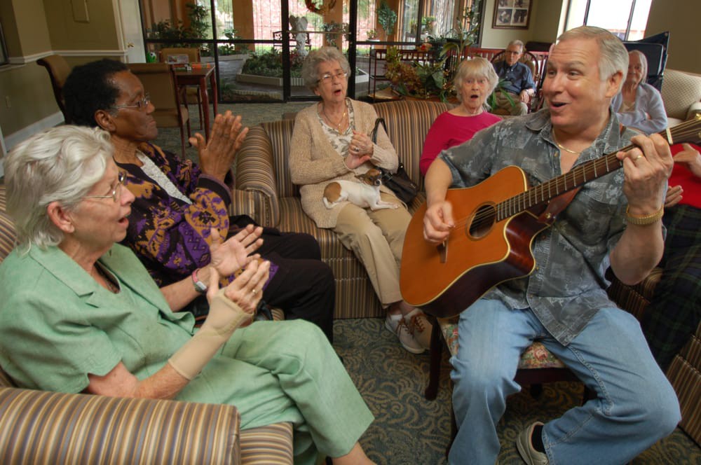 A group of elderly people enjoying music, with one playing the guitar and others clapping along.