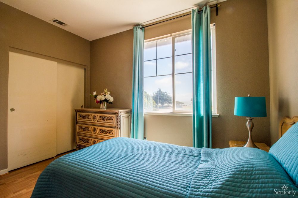 A cozy bedroom with blue curtains and bedding, featuring a wooden dresser and a bedside lamp.