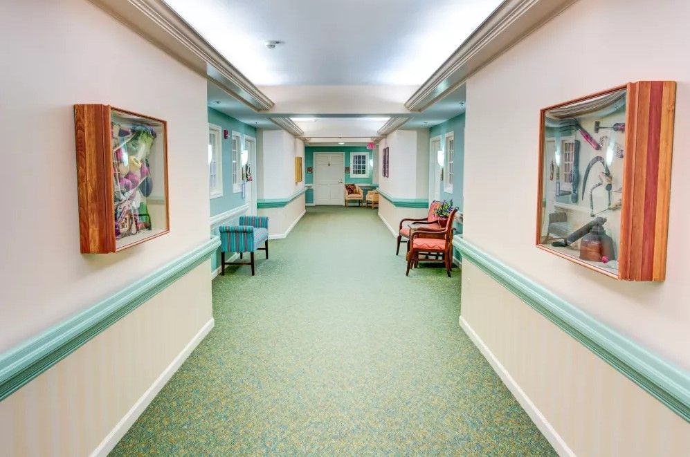 A well-lit hallway with carpeted flooring, adorned with chairs and paintings on the walls.