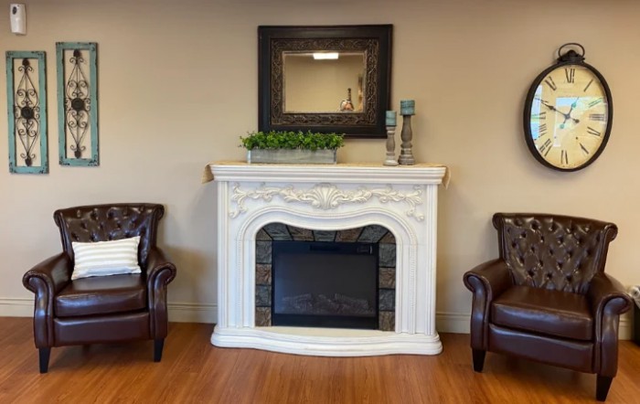 A cozy living room setup featuring a decorative white fireplace flanked by leather armchairs, with a mirror and candles on the mantelpiece.