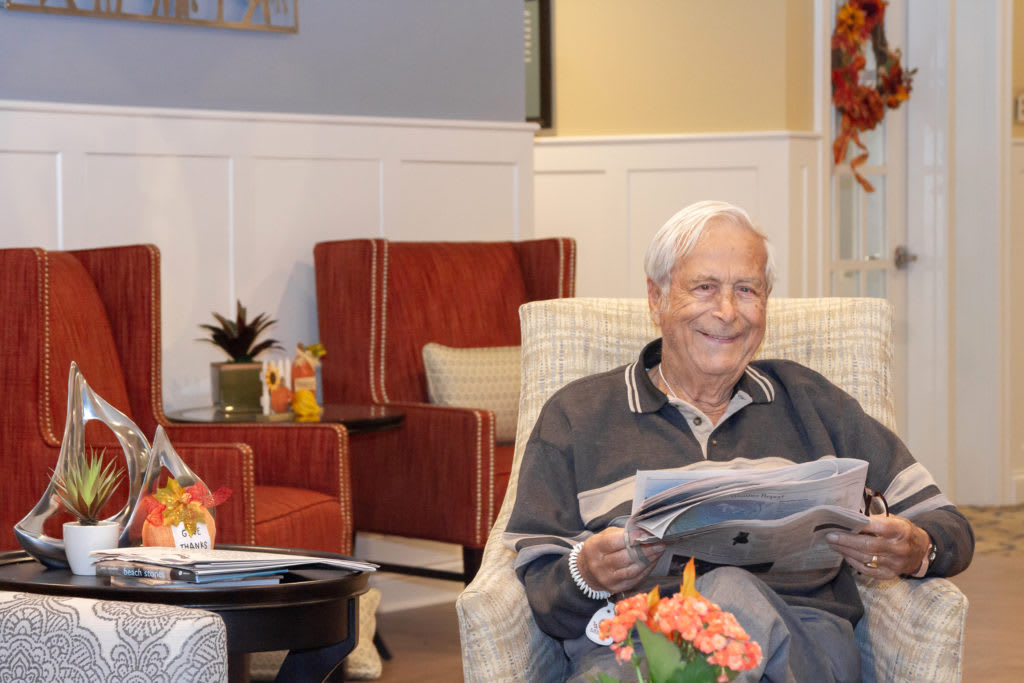 An elderly man sits comfortably in a living room, smiling as he reads a newspaper.