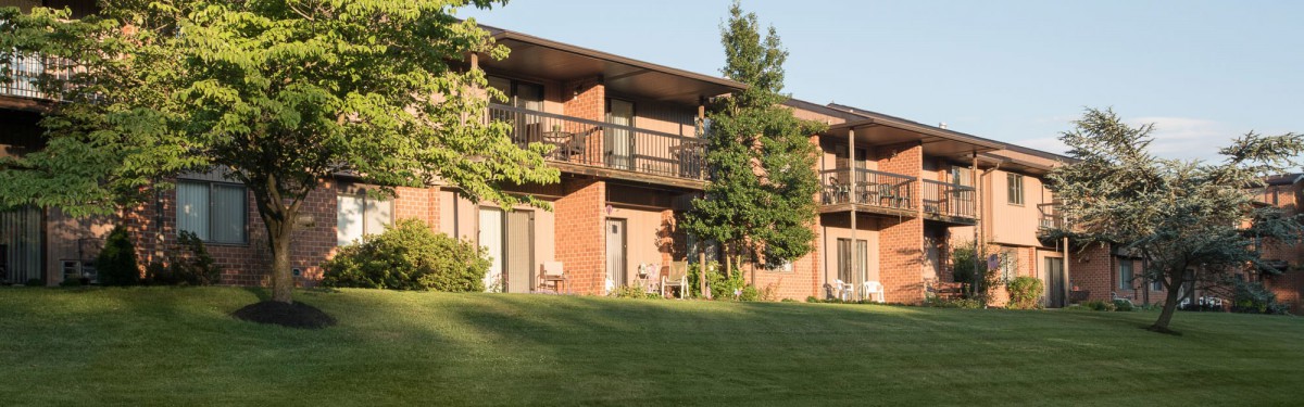 A two-story apartment building with brick exterior and balconies, surrounded by a well-kept lawn and trees.