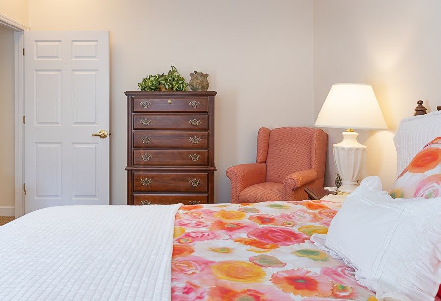 A cozy bedroom featuring a wooden dresser, a colorful bedspread, an armchair, and a lamp for warm lighting.