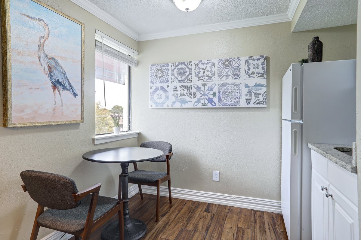 A small kitchen dining area with a round table, two chairs, artwork on the walls, and a refrigerator in the corner.