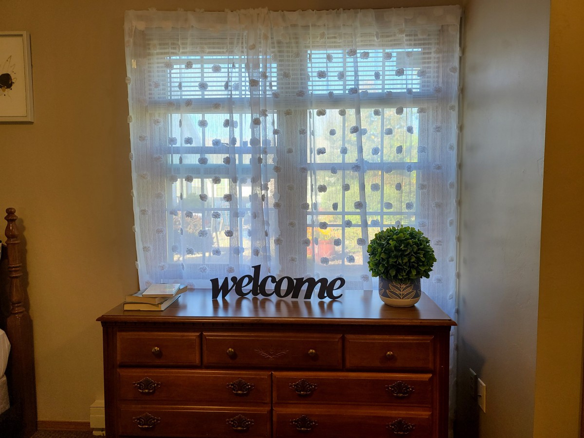 A cozy room with a dresser featuring a welcome sign and a small plant by the window.