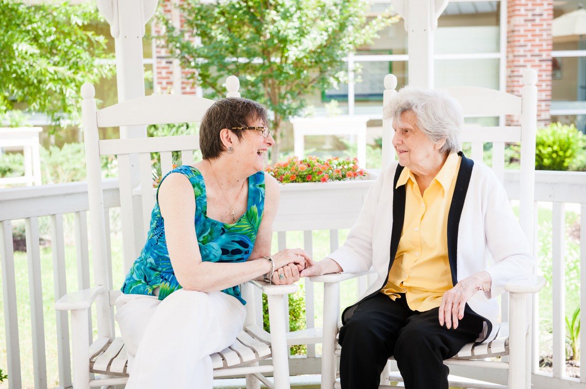 Two women sitting outdoors, smiling and holding hands in conversation.