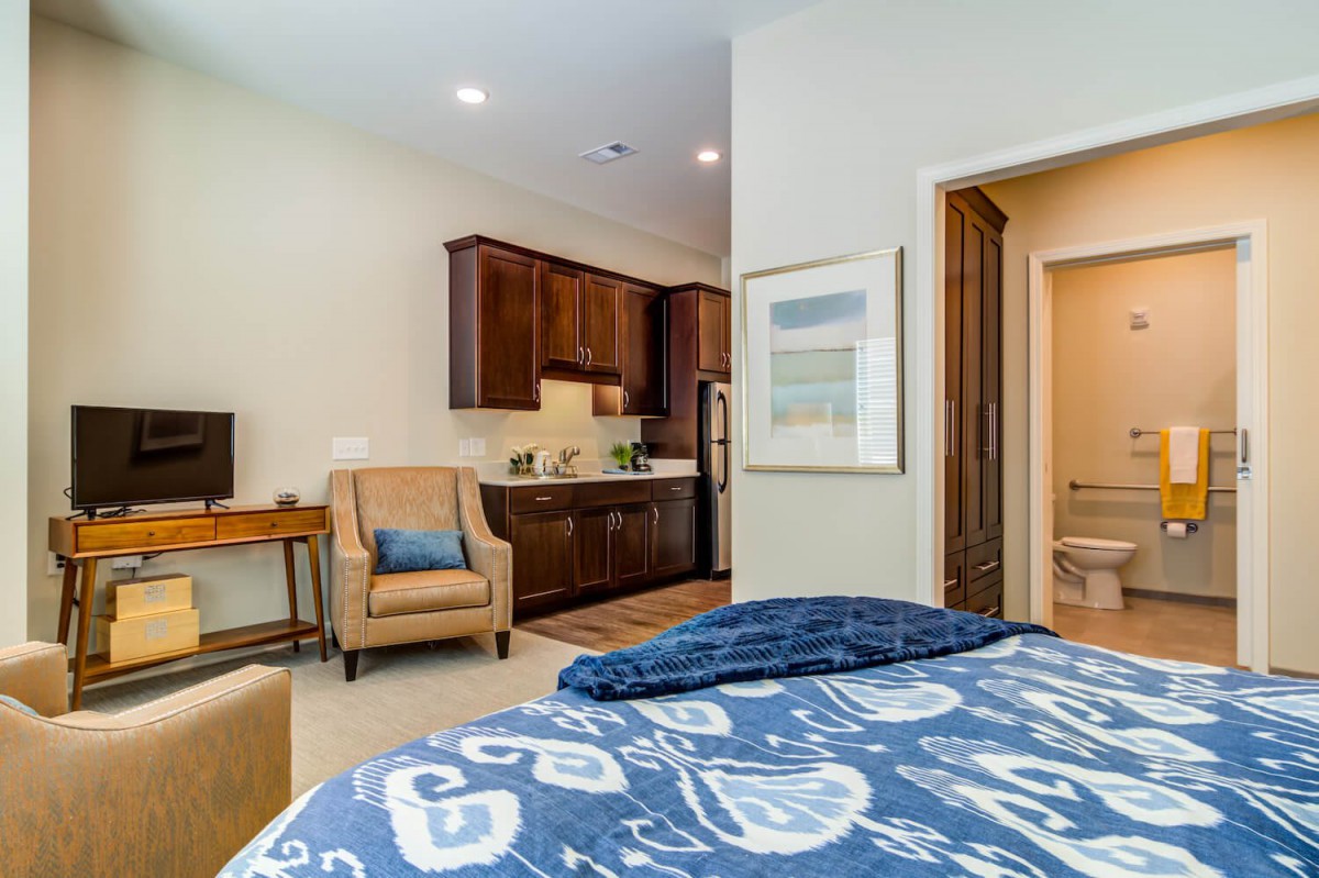 A cozy bedroom with a blue patterned blanket, featuring a small kitchenette and an armchair.
