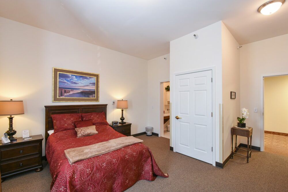 A cozy bedroom featuring a red bedspread, warm lighting from a table lamp, and wall art above the bed.
