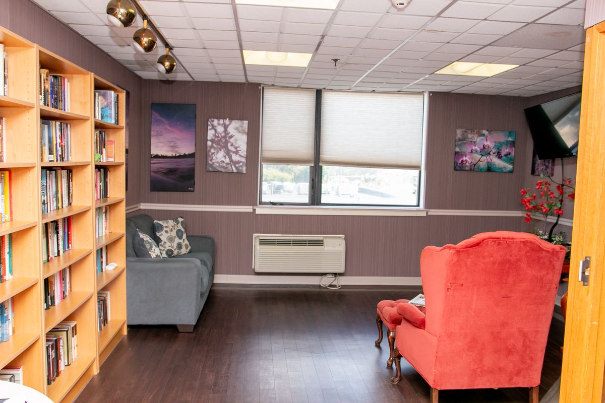 A cozy living room with a bookshelf, armchair, and sofa alongside artwork and a window.