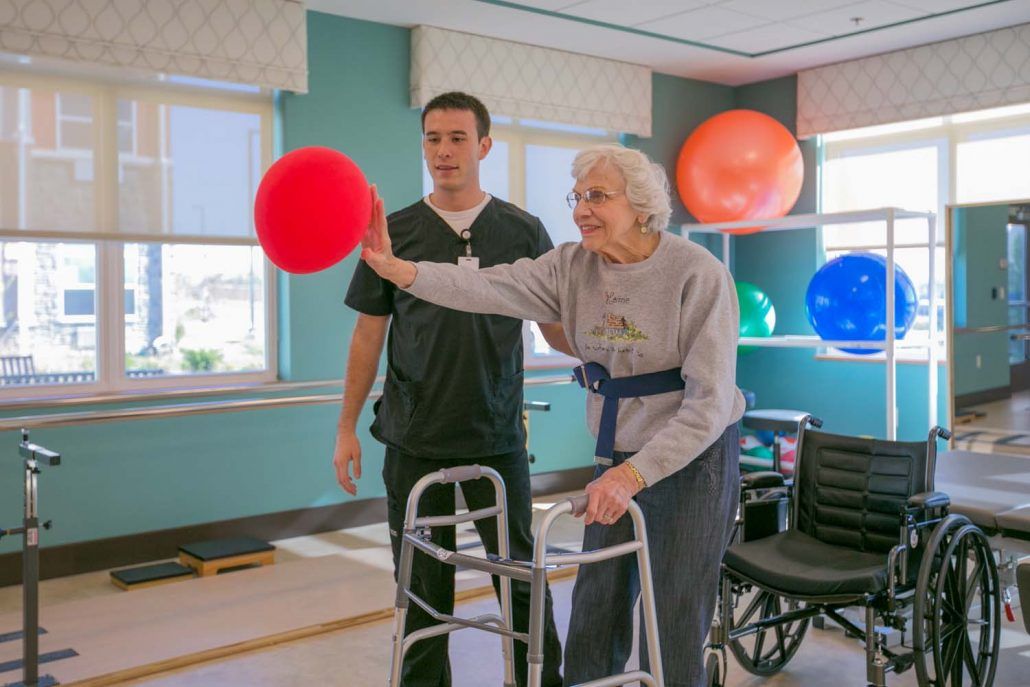 An elderly woman participates in a physical therapy session with the assistance of a healthcare professional, using a walker and engaging with a red ball.