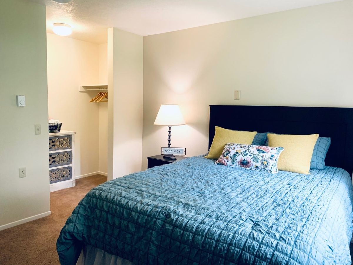 A cozy bedroom with a neatly made bed, a bedside lamp, and an open closet.
