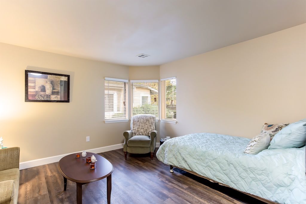 A cozy bedroom with a bed, armchair, and a small table, featuring wooden flooring and a large window.