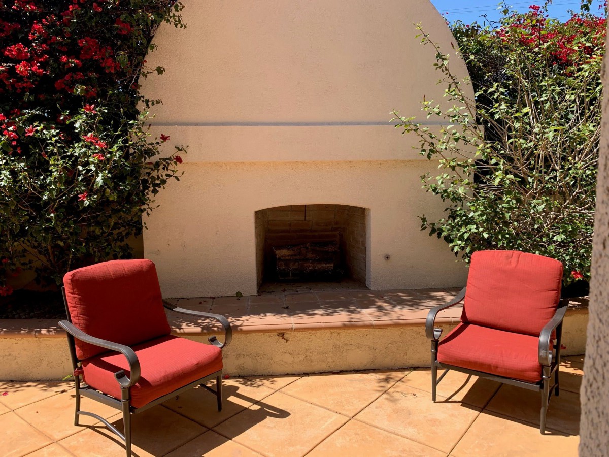 A sunny outdoor patio with a built-in fireplace and two red cushioned chairs, surrounded by lush greenery.