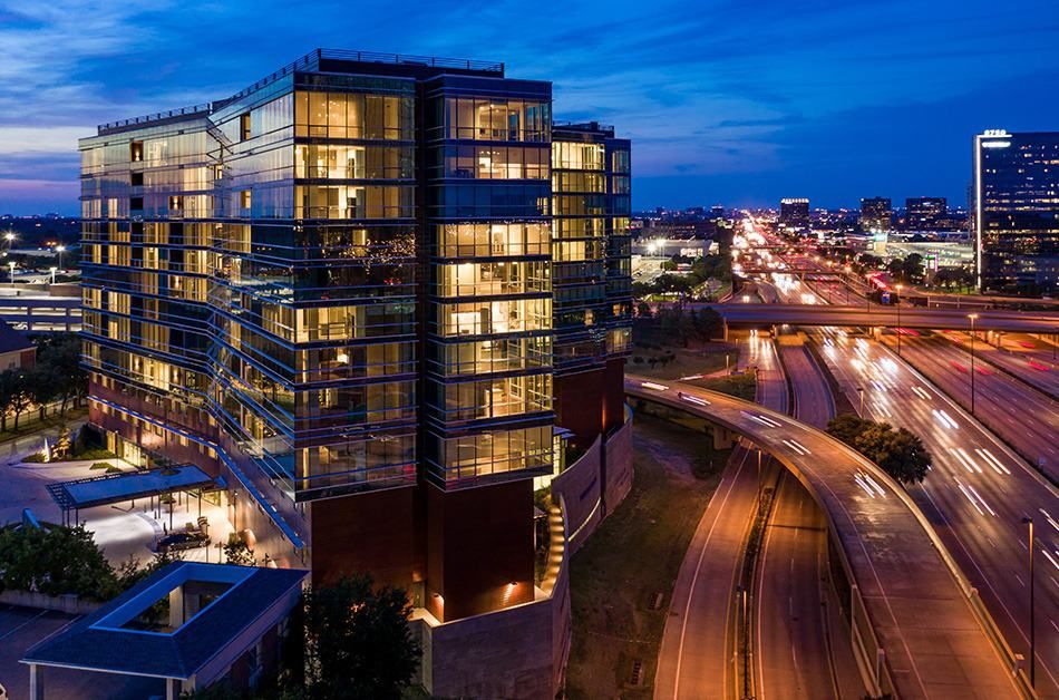 Modern glass building illuminated at night with city traffic in the background.