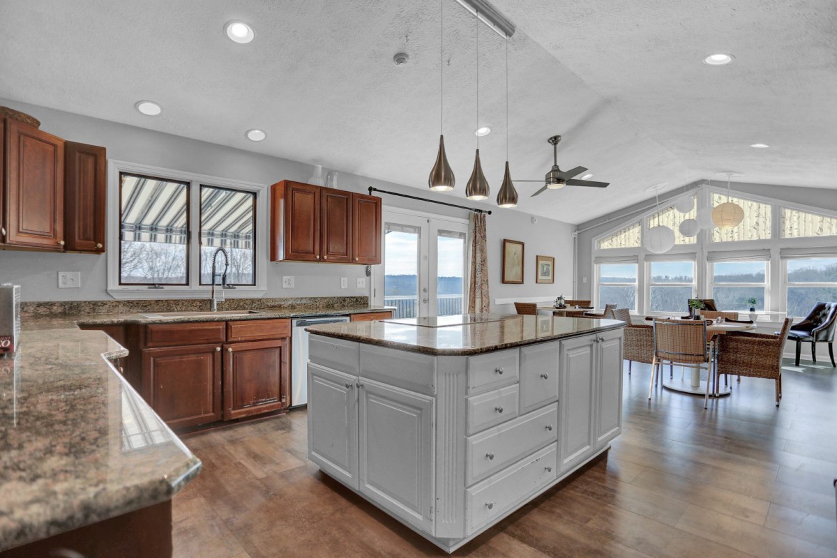 A modern kitchen with an island, featuring pendant lights and wooden cabinets, opens into a living area with large windows.