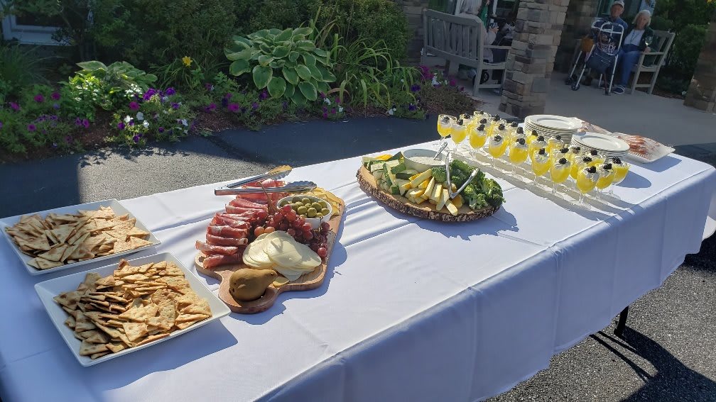 A sunny outdoor setting with a table displaying charcuterie, cheese, fruits, and drinks.