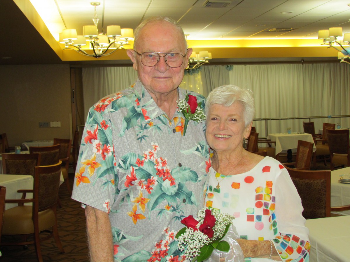 An elderly couple smiling happily, with the man wearing a floral shirt and the woman holding flowers.