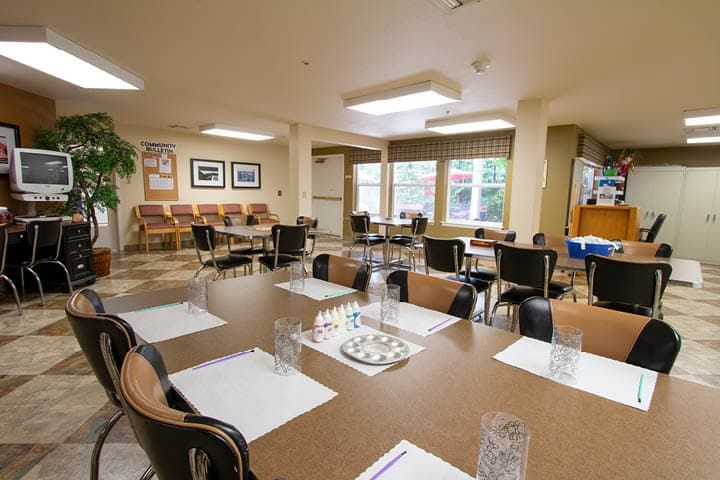A well-lit dining room with multiple tables and chairs arranged neatly, featuring place settings and large windows.