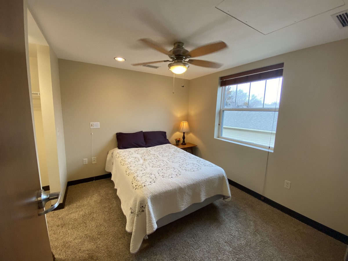 A cozy bedroom featuring a bed with white linens, a bedside lamp, and a ceiling fan.