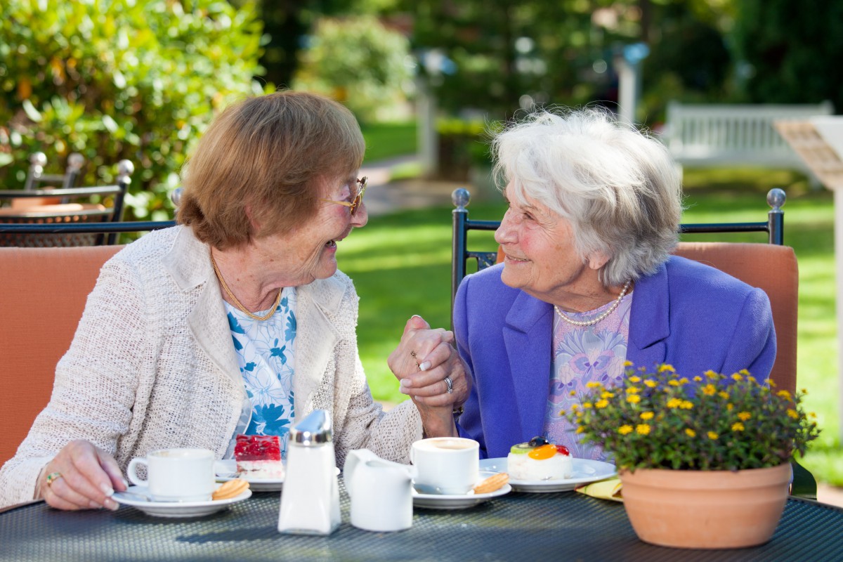 Two elderly women enjoying a conversation and coffee in a garden setting.