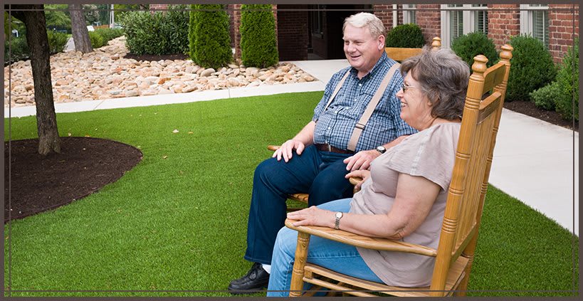 An elderly couple sitting outdoors on wooden chairs, enjoying a relaxing time in the garden.
