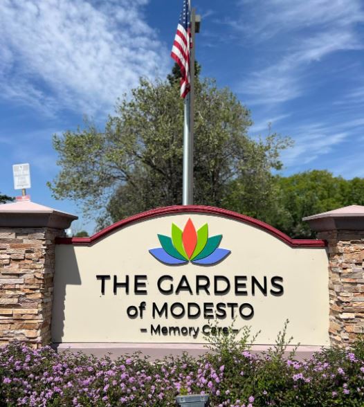 A sign for The Gardens of Modesto Memory Care facility, featuring landscaping and an American flag.