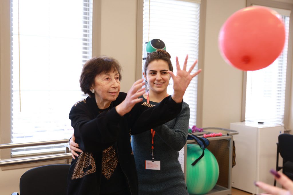 An elderly woman engaging in a physical therapy session with a therapist, using a red ball for exercise.