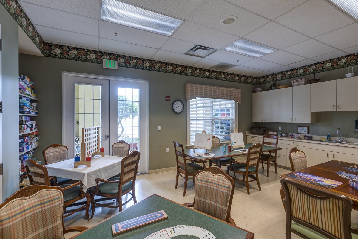 A cozy dining room with multiple tables and chairs, adjacent to a kitchen area, well-lit by natural light from the windows.