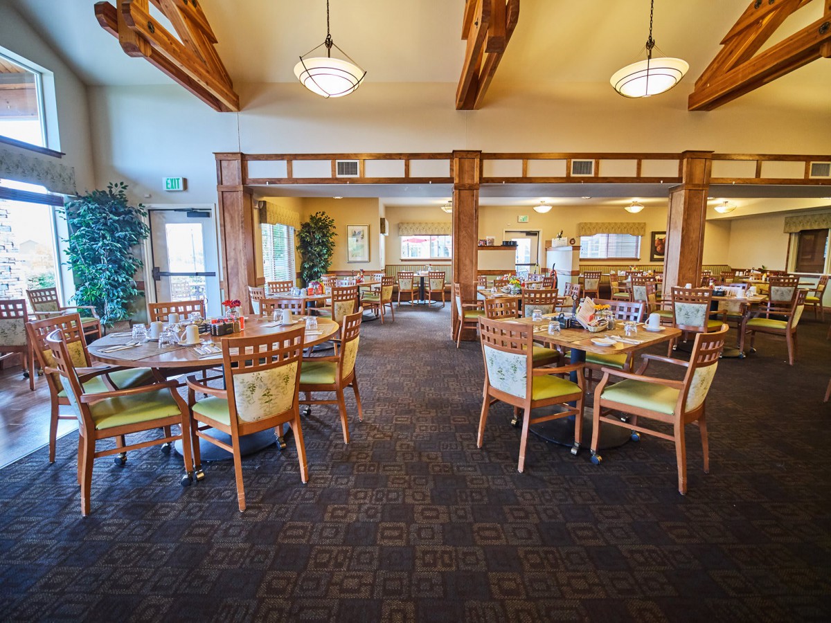 A spacious dining area with wooden beams and chandeliers, featuring tables and chairs arranged on a carpeted floor.