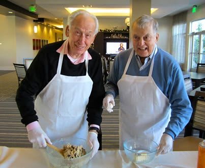 Two elderly men wearing aprons are engaged in a cooking activity together.