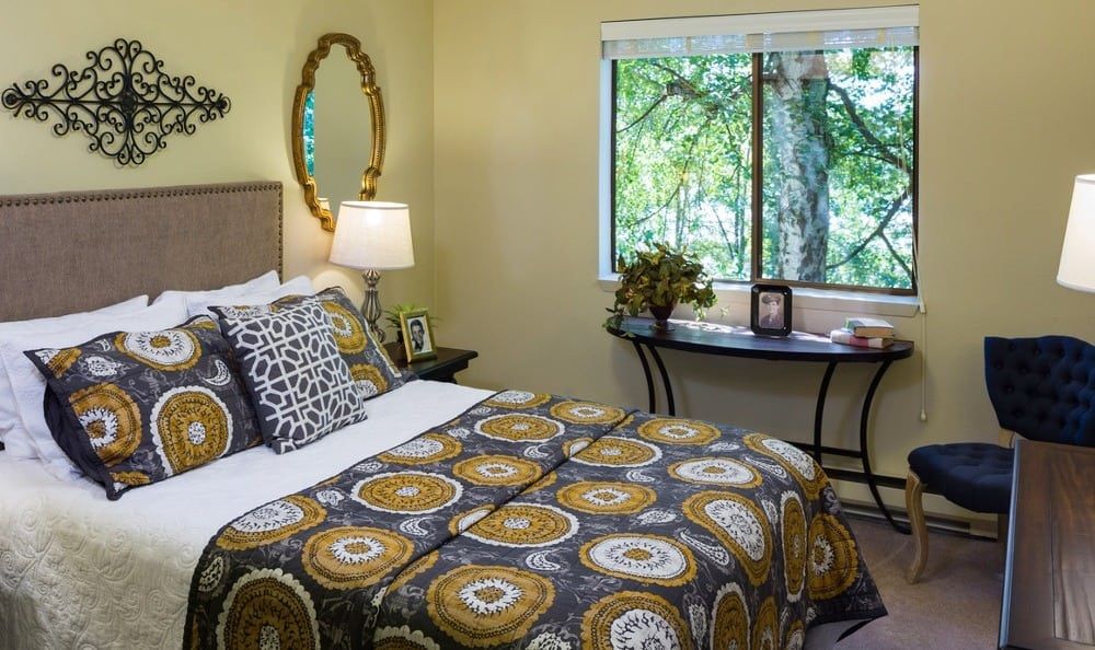 A cozy bedroom features patterned bedding, a decorative mirror, and a window with a view of greenery.