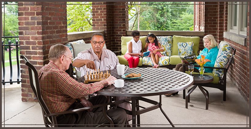 Family relaxing on a porch, with two men playing chess and a mother and daughter sitting on a couch.