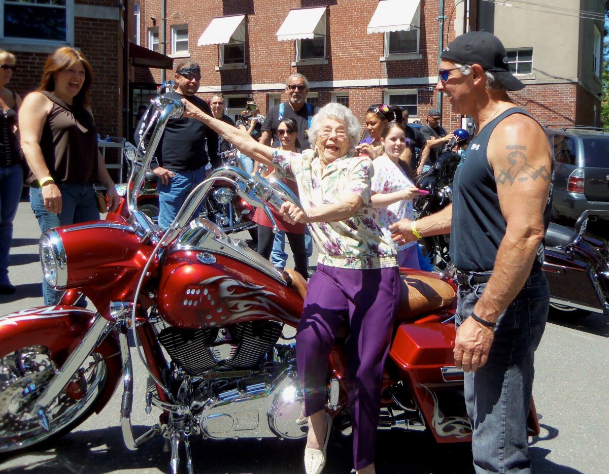 An elderly woman joyfully poses on a motorcycle during a community event, surrounded by onlookers.