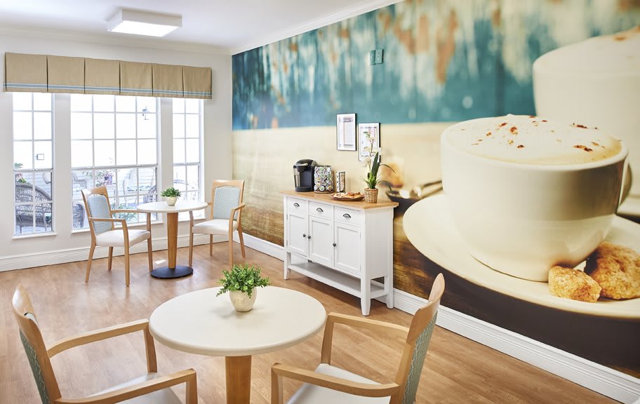 A cozy cafe interior with wooden tables and chairs, a large mural on the wall, and a coffee station set up beside a window.