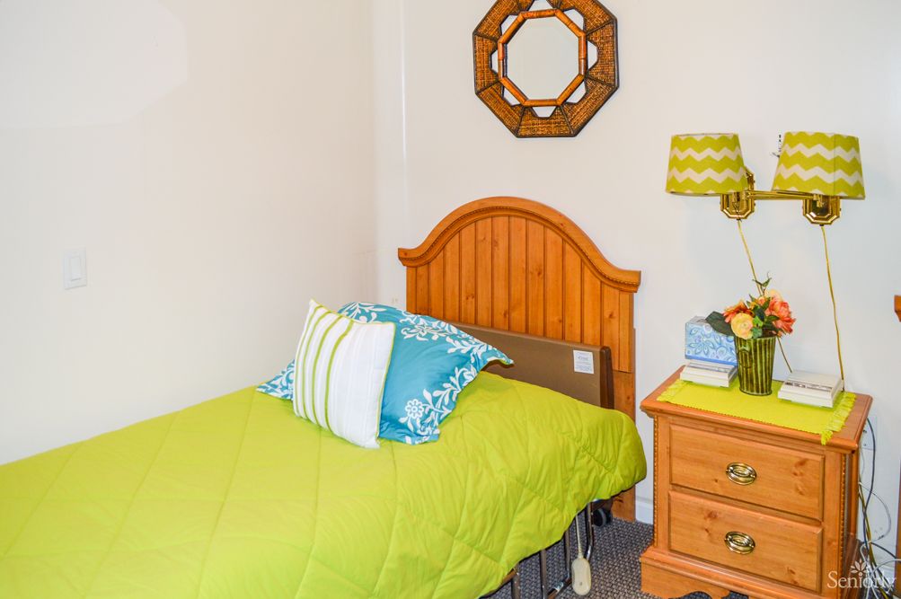 A cozy bedroom featuring a single bed with green bedding, a wooden headboard, and a decorative mirror above the bed. A nightstand with a lamp and some decor items completes the space.