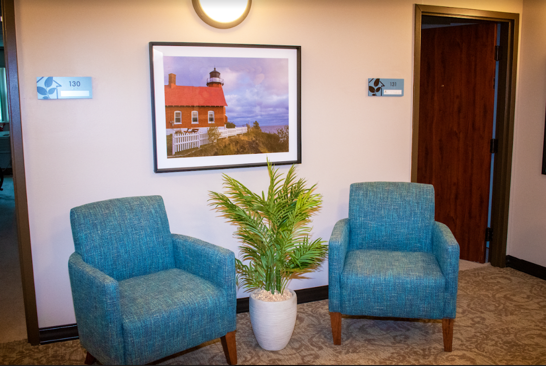 A cozy waiting area featuring two blue chairs, a potted plant, and a framed lighthouse photograph on the wall.