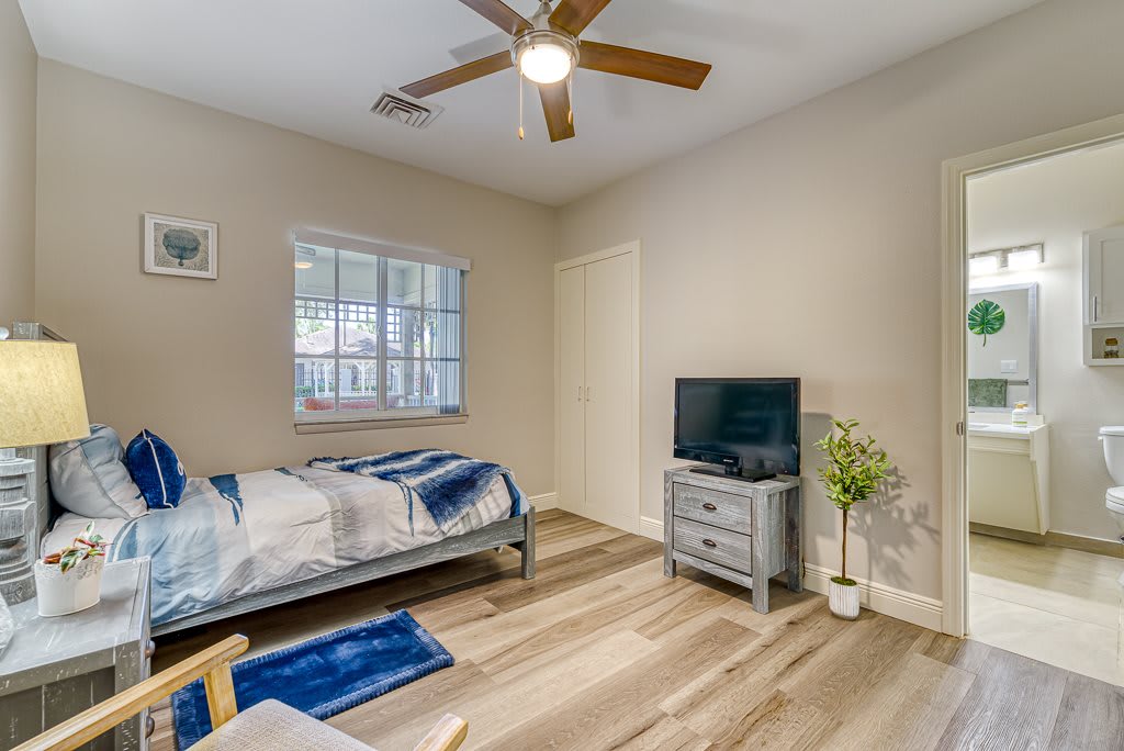 A neatly arranged bedroom featuring a single bed, a television on a nightstand, and a ceiling fan.